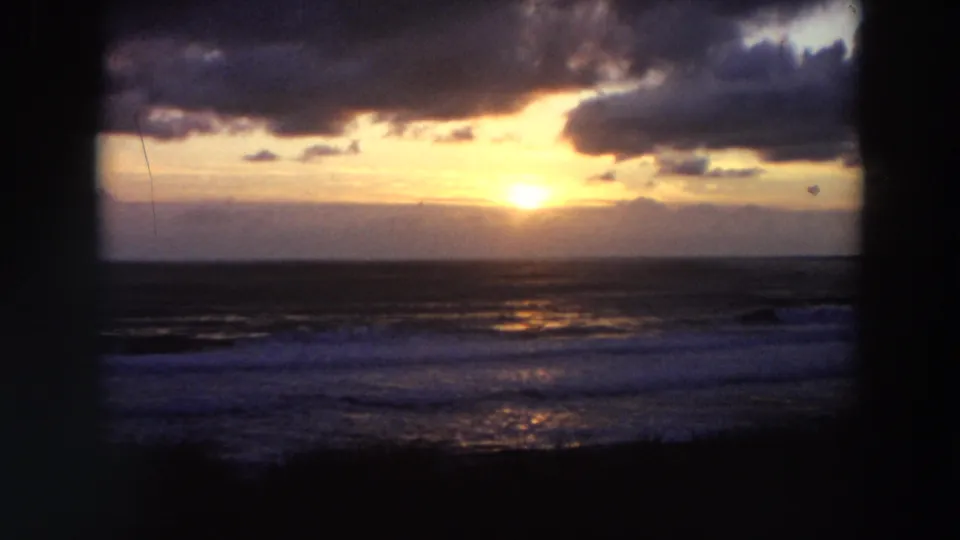 1964 California 8mm frame showing a low sun over the ocean, cloud bands, surf, and dark framing silhouettes.
