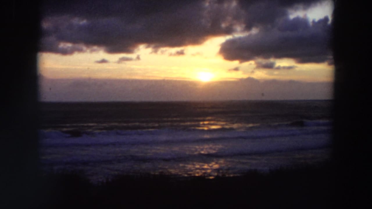 1964 California coast view with the sun glowing above the horizon, surf below, and dark clouds across the sky.