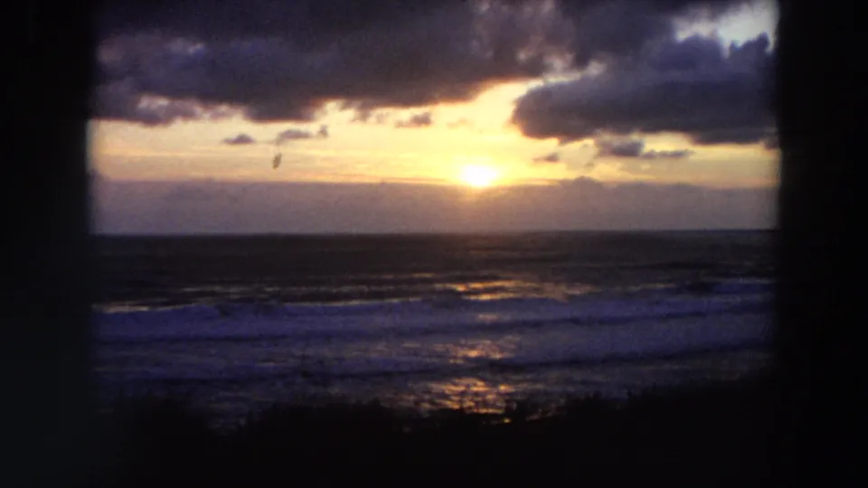 Vintage 8mm coastal sunset frame from California, showing clouded sky, ocean surf, and warm reflected light.
