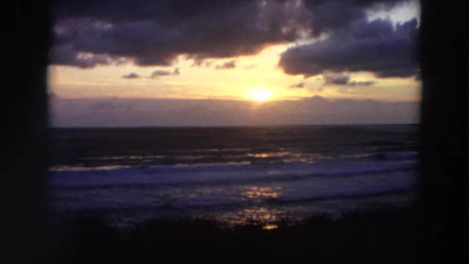 1964 California 8mm view of a coastal sunset, with dark foreground framing, heavy clouds, and reflective surf.