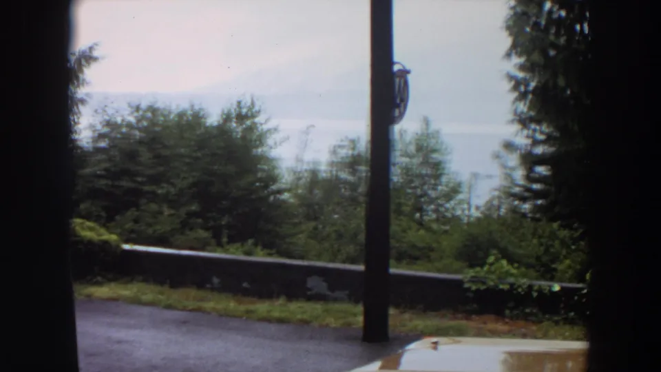 1964 California 8mm frame showing a sheltered view toward trees, low wall, wet pavement, and pale distant sky or water haze.