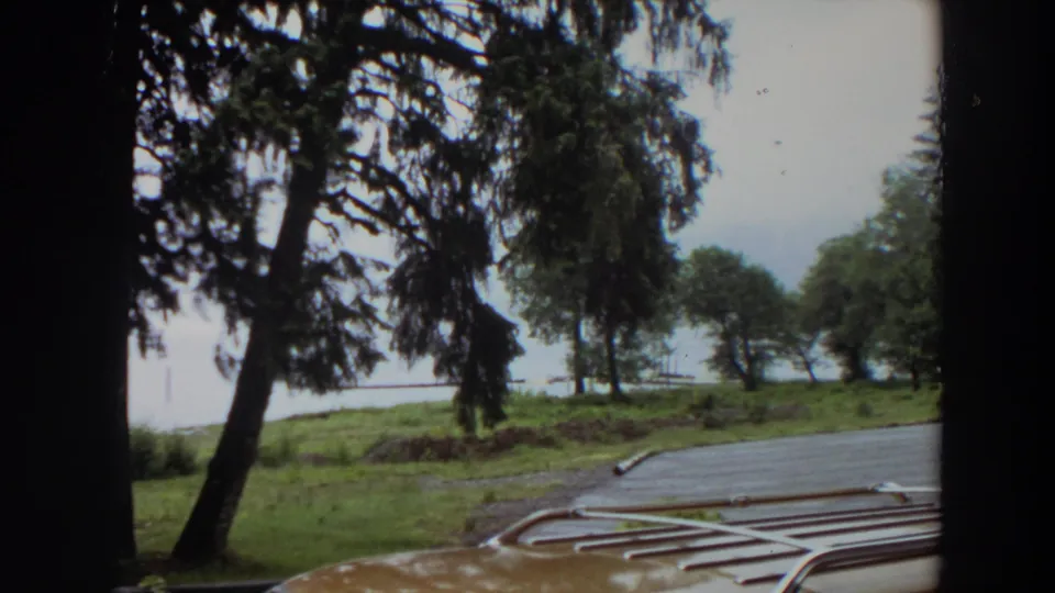 1964 California 8mm frame looking over a car roof rack toward wet asphalt, green trees, grass, and a calm body of water.