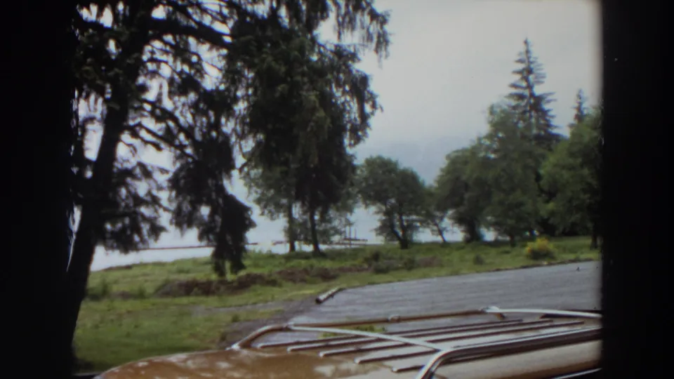 1964 California 8mm frame with the car roof rack low in view and a damp wooded parking area beside water.