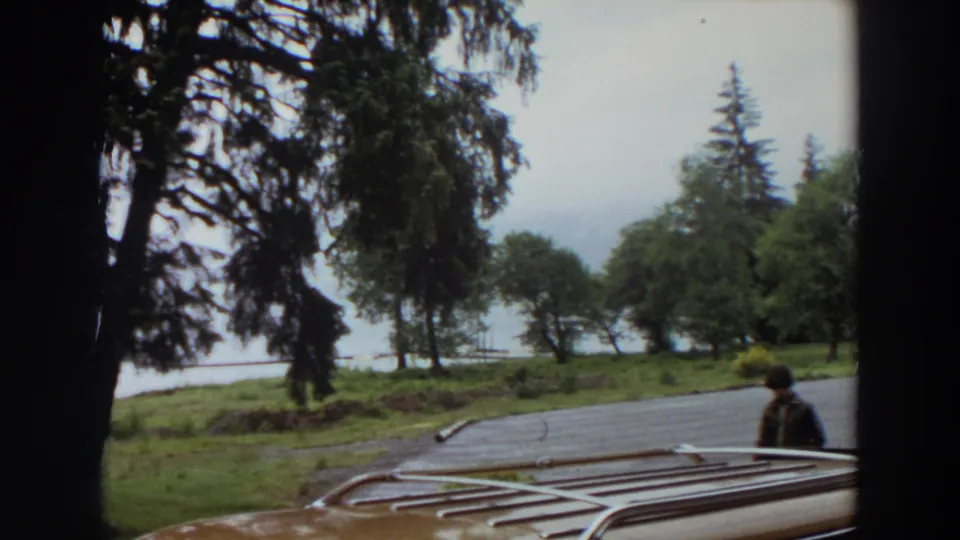 The final 1964 California frame holds on wet asphalt, a car roof rack, trees, grass, and a misty waterline beyond.