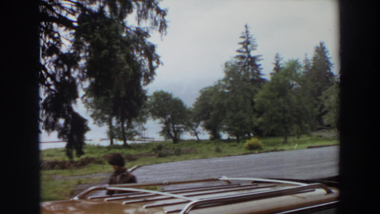 1964 California home-movie frame showing a person walking beside a car roof rack near trees and a lakeside lot.