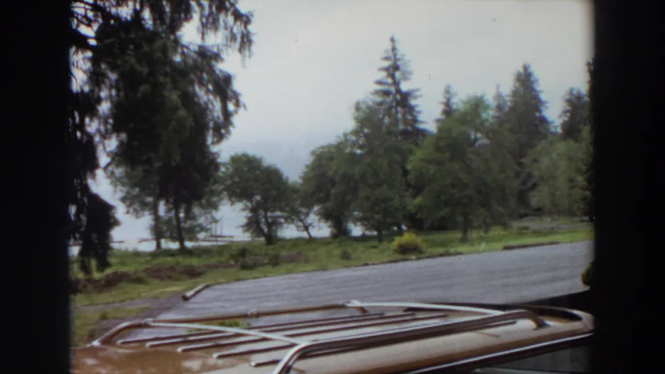 1964 California 8mm frame looking across a car roof rack toward a paved lakeside lot and dense trees.