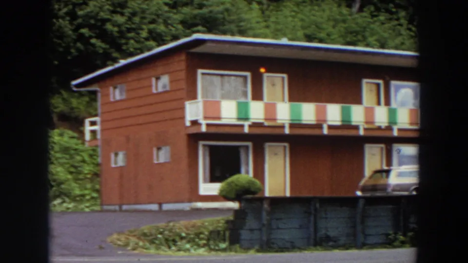 1964 California home-movie view of a two-story wood-sided motel with colorful railings and a parked car.
