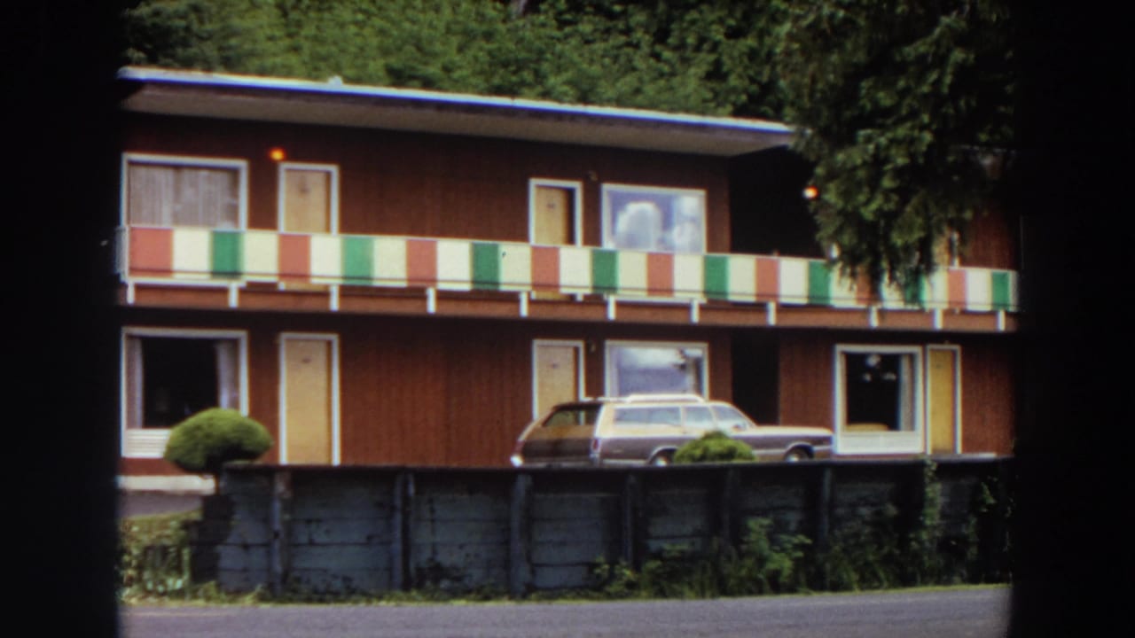 Red motel exterior with balcony panels, parked station wagon, shrubs, and trees.