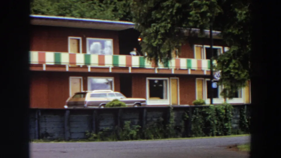 1964 California frame showing the motel from a slightly shifted angle, with striped balcony railing, a parked wagon, trees, and a small distant person.