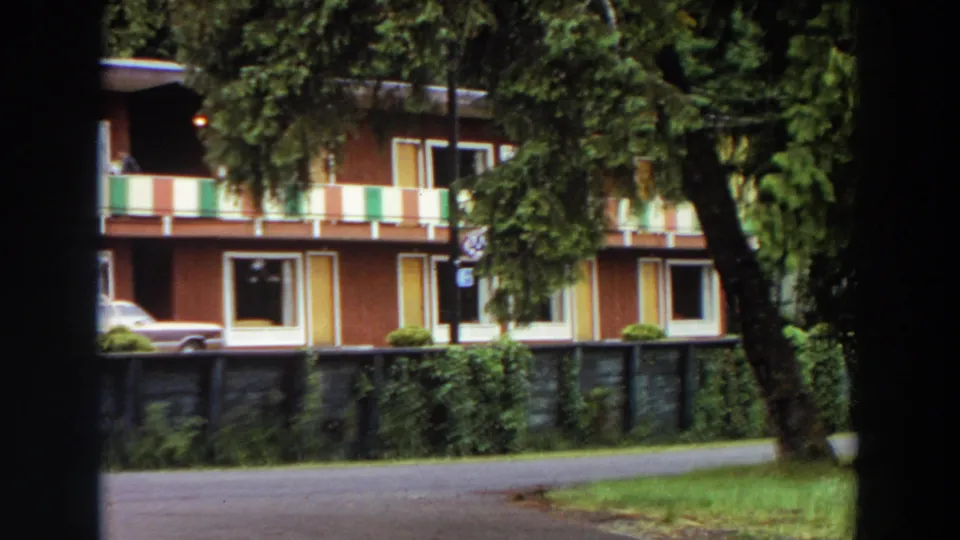1964 California 8mm frame of the red motel partly obscured by trees, with striped balcony panels, room doors, shrubs, and road foreground.