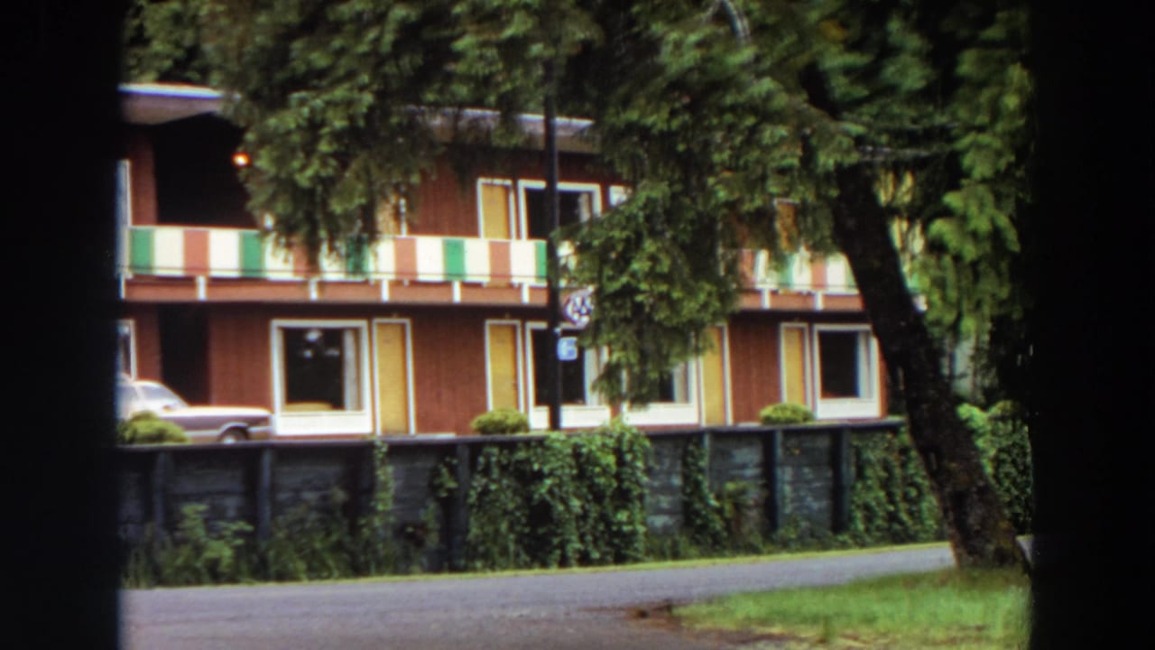 Two-story apartment building behind trees with a parked station wagon and low fence.