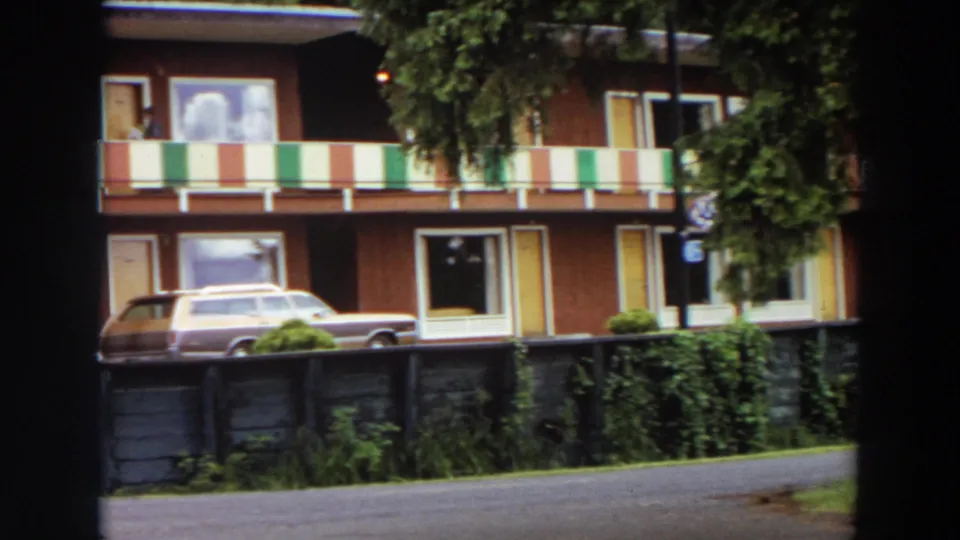 1964 California frame of a two-story apartment exterior with striped balcony panels, shrubs, a low fence, and a brown-and-beige station wagon.