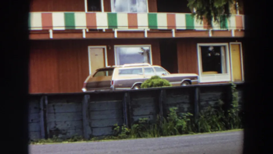 Closer 1964 California 8mm view of the apartment exterior, striped balcony, parked station wagon, fence, shrubs, and paved foreground.