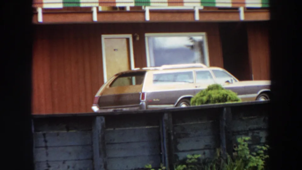 1964 California frame with a brown-and-beige station wagon, apartment windows and doors, striped balcony edge, fence, and greenery.
