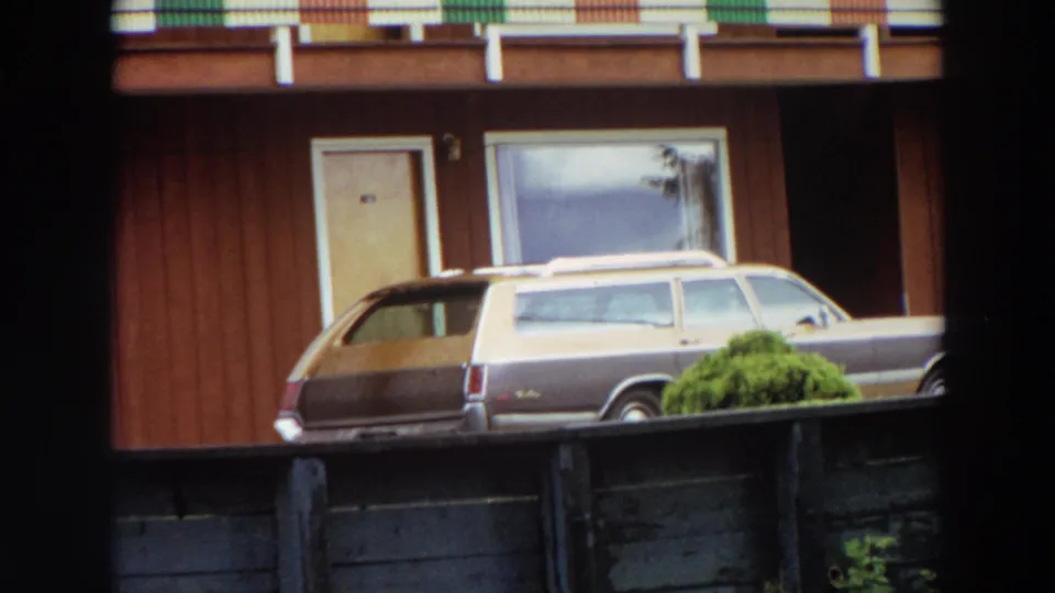 1964 California 8mm frame showing a two-tone station wagon parked outside a wood-sided house, partly framed by a dark foreground edge.