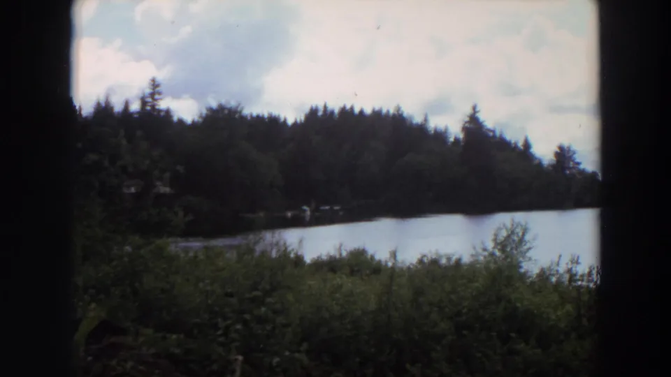 1964 California lakeside view on 8mm film, with still water, dense evergreen trees, clouds, and dark frame edges from the shooting position.