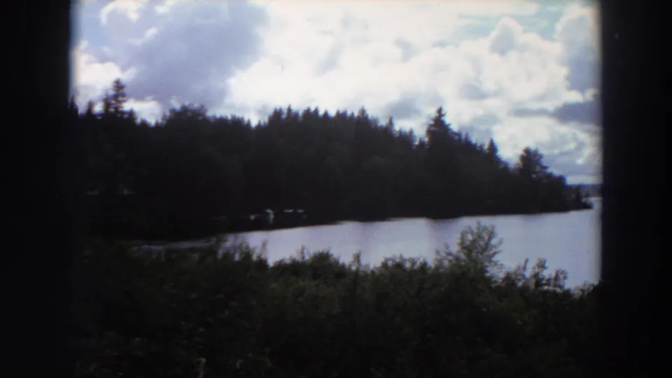1964 California home-movie frame of a lake bordered by dark evergreen trees, with foreground brush and a bright cloud-filled sky.
