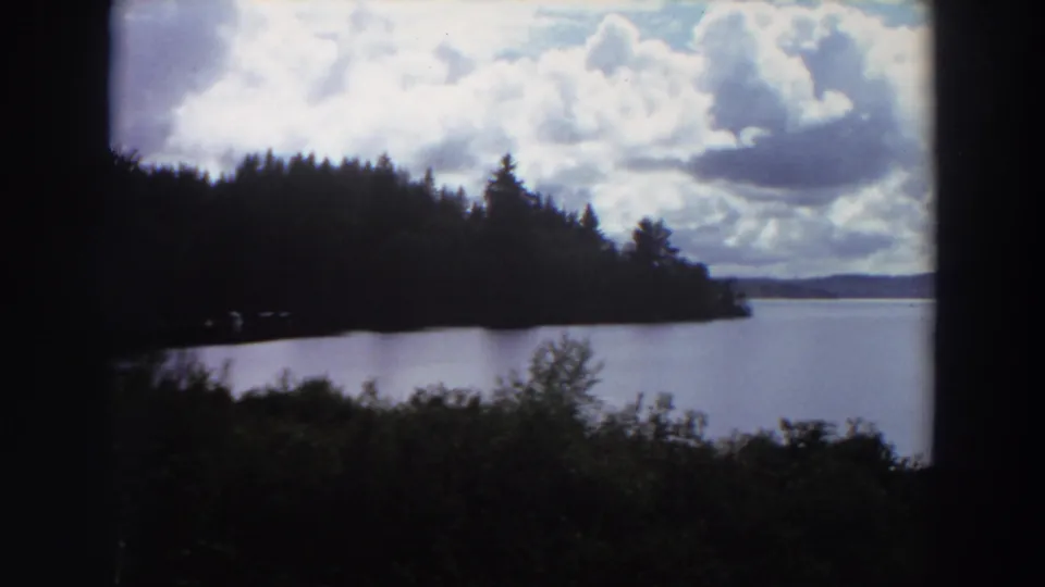 1964 California 8mm view across a lake toward a dark tree-lined shore and distant hills beneath a bright cloud-filled sky.