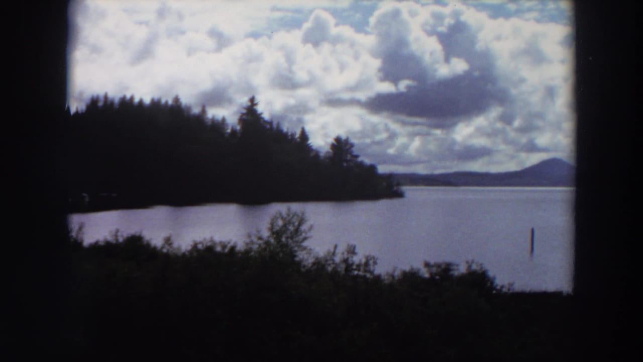 Wide lake view with forested point, distant mountain, clouds, and a post in the water.