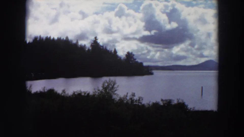 1964 California 8mm frame showing calm lake water, a wooded point, distant mountain profile, dramatic clouds, and a narrow post in the water.