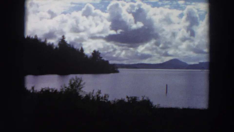 1964 California lake scene on 8mm film, with dark trees at left, open water, distant mountains, large clouds, and a solitary post.