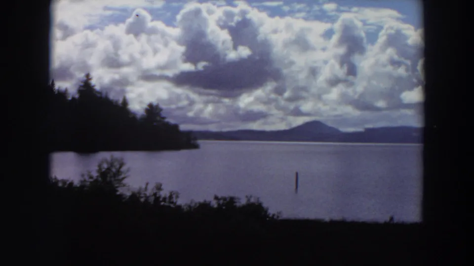 1964 California 8mm landscape looking over a lake from shaded foliage toward silhouetted trees, distant hills, and high clouds.