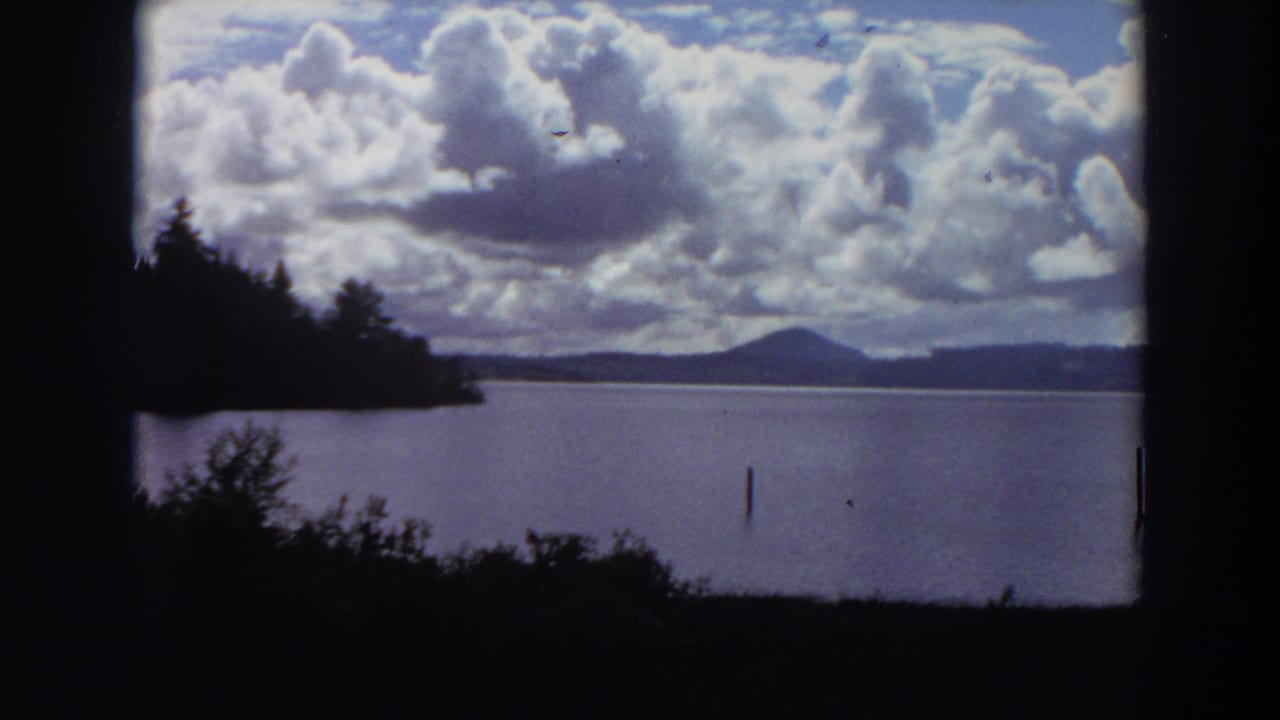 Calm lake with dark trees, distant hills, and large clouds overhead.