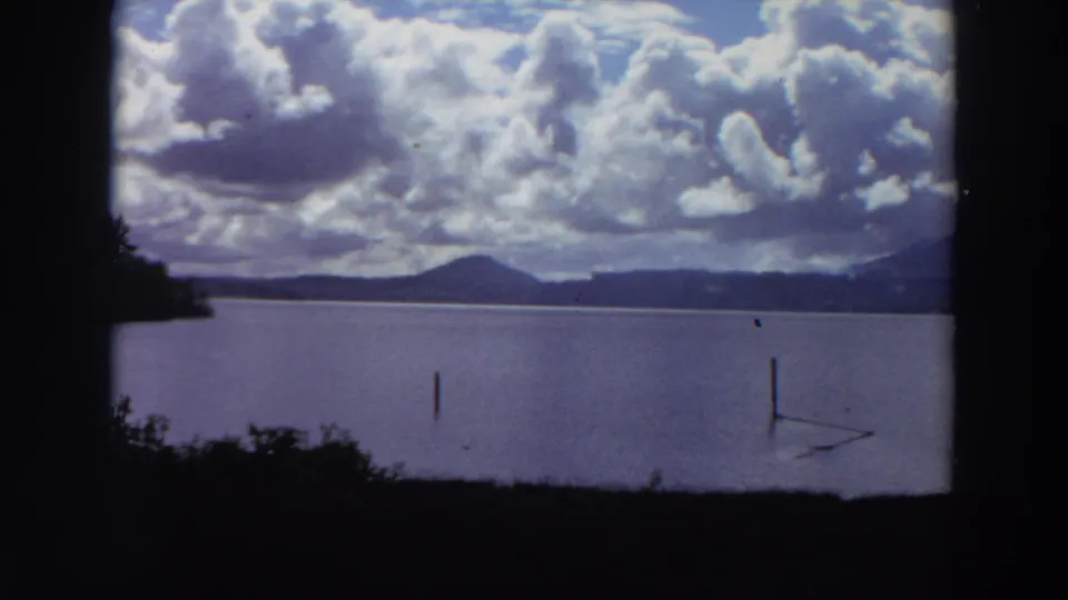 1964 California 8mm frame showing a broad lake, a dark wooded shore, distant mountains, and cumulus clouds across the sky.
