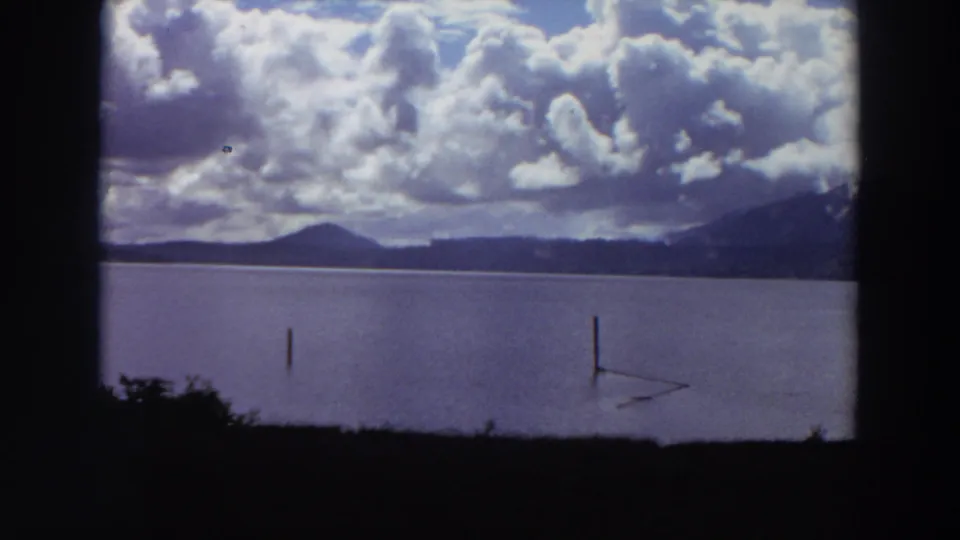 1964 California lakeshore frame with still water, two posts, a low mountain ridge, and dark foreground vegetation under large clouds.