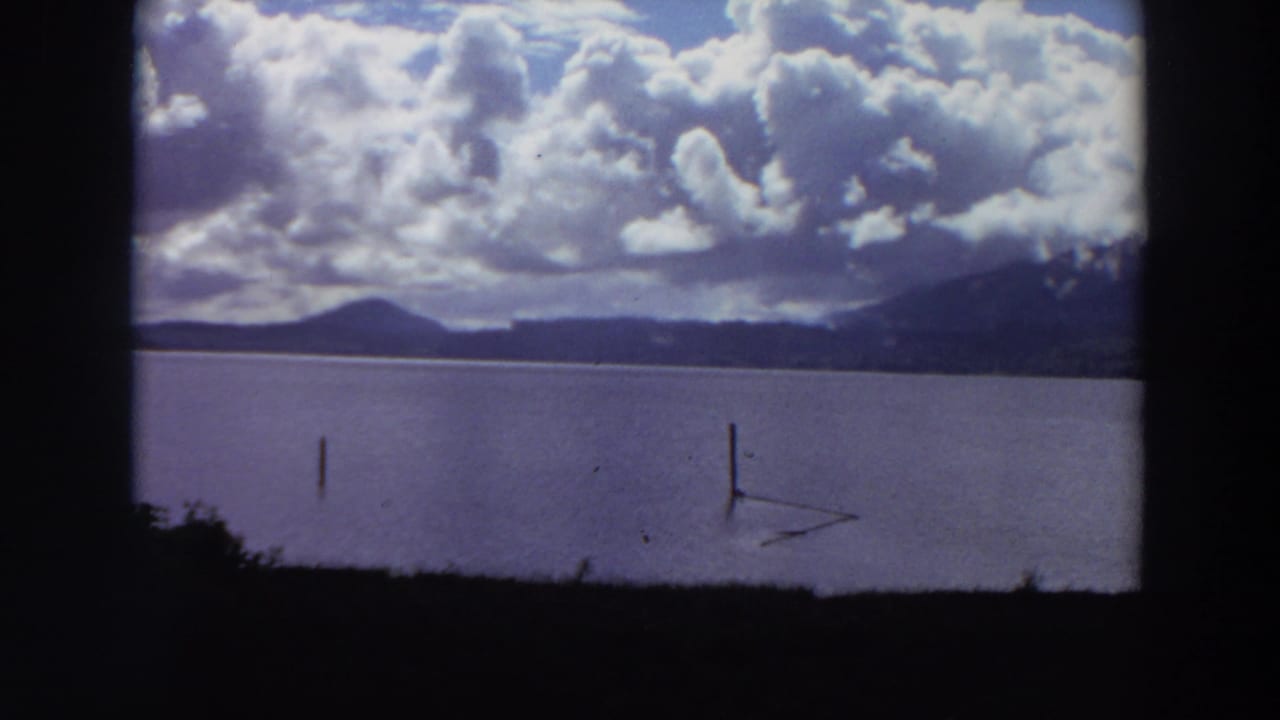 1964 California 8mm view of a lake with wooden posts in the water, mountain ridges beyond, and large cumulus clouds overhead.
