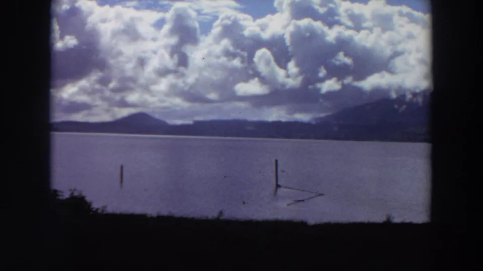 1964 California 8mm view of a lake with wooden posts in the water, mountain ridges beyond, and large cumulus clouds overhead.