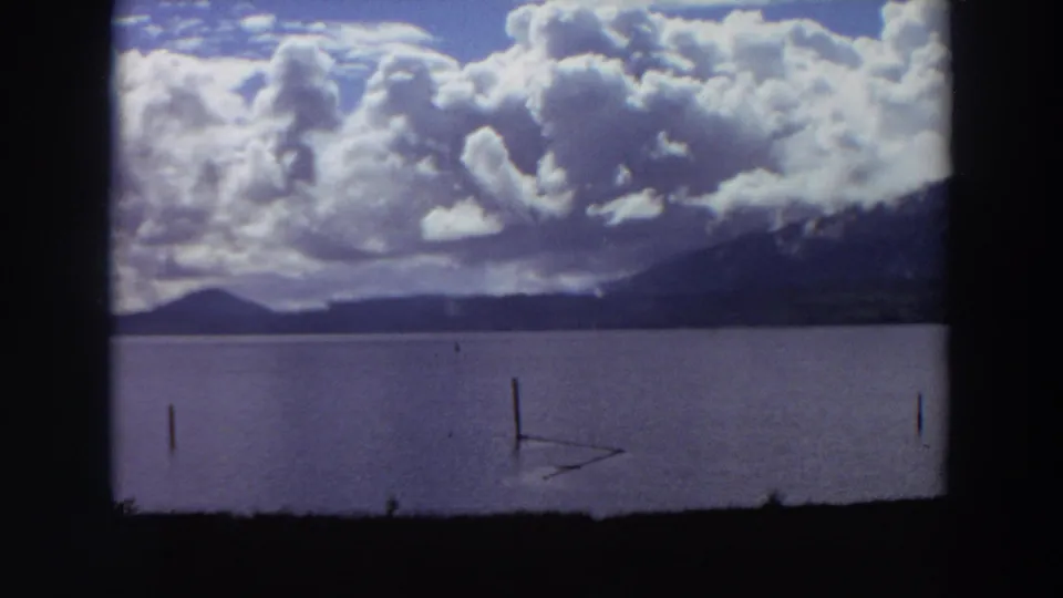 1964 California lakeside scene with still water, scattered posts, distant mountains, and dramatic cloud cover within the film frame.