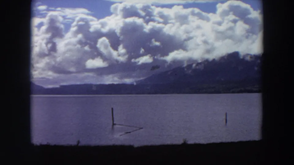 1964 California home-movie frame showing wooden posts in a lake, a mountain ridge, and bright cumulus clouds across the sky.