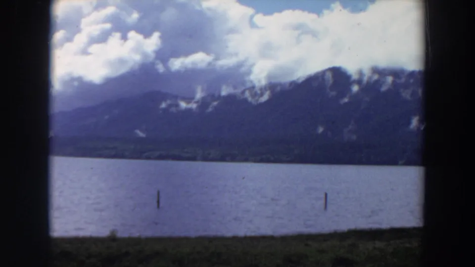 1964 California 8mm landscape with a lake foreground, two water posts, dark mountain slopes, and clouds hanging over the ridgeline.