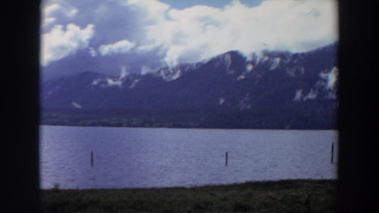 1964 California 8mm frame showing a lakeside view with wooden posts, grassy shore, distant mountains, and bright clouds.