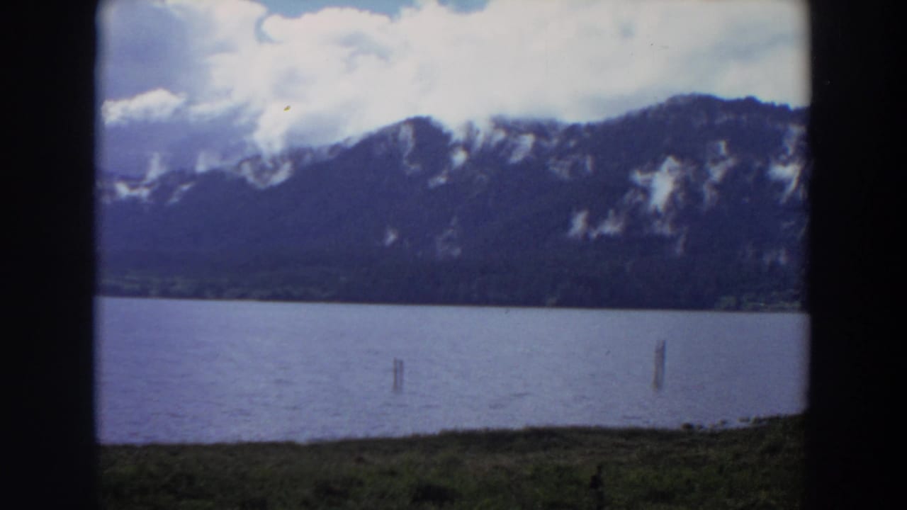 Lake surface with wooden posts, distant mountain ridge, and clouds across a bright sky.