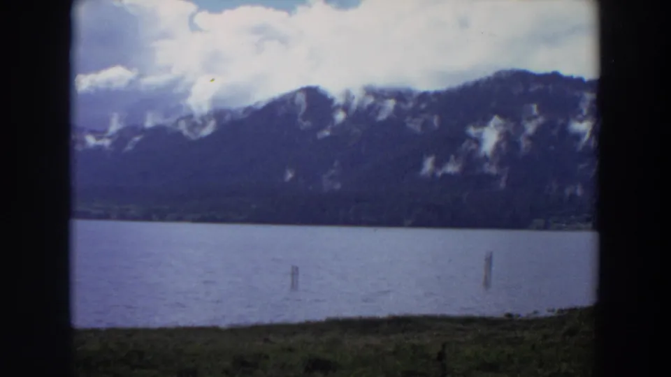 1964 California 8mm view across a lake toward a tree-covered mountain ridge under white clouds.