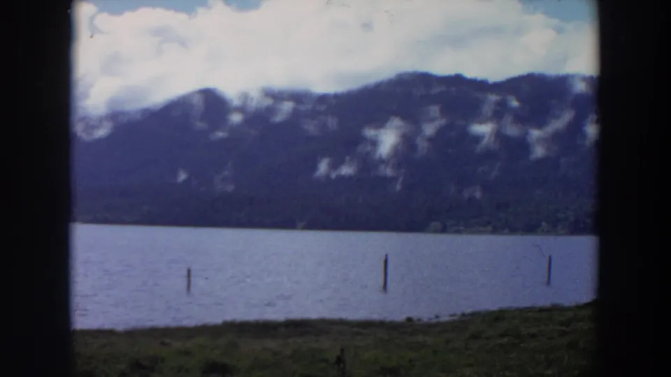 1964 California 8mm frame with lake posts, a grassy edge, and a dark mountain ridge partly marked by pale snow or rock.