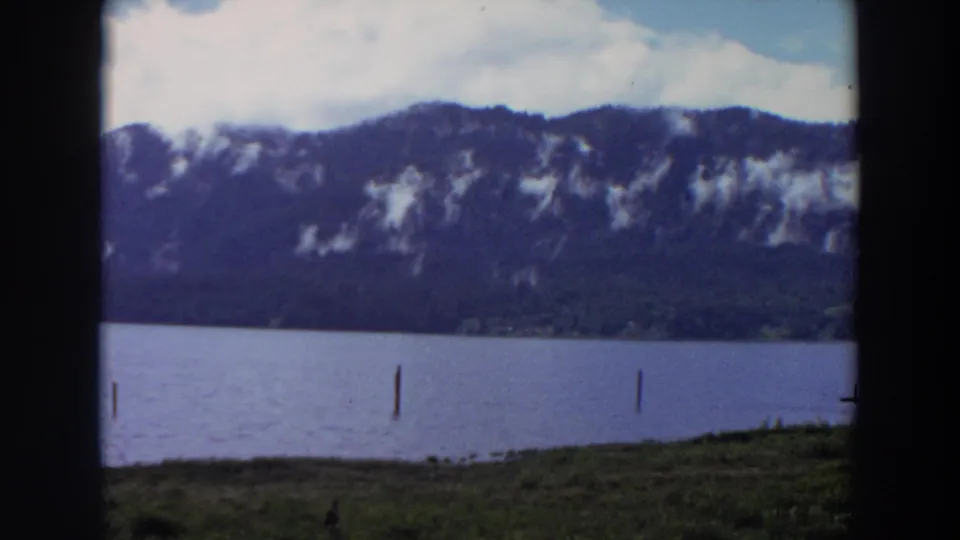 1964 California 8mm frame showing a calm lake, grassy shoreline, wooden posts, and mountains beneath bright cloud cover.