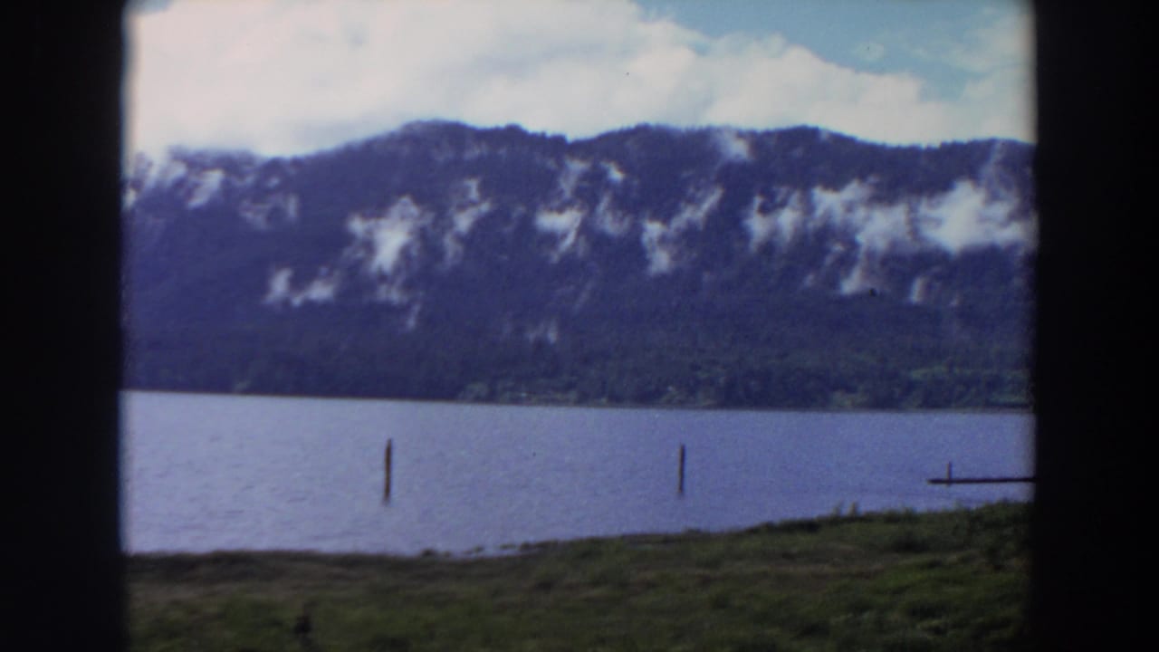 1964 California 8mm frame showing a calm lake, grassy shore, mountain ridge, and scattered low clouds.