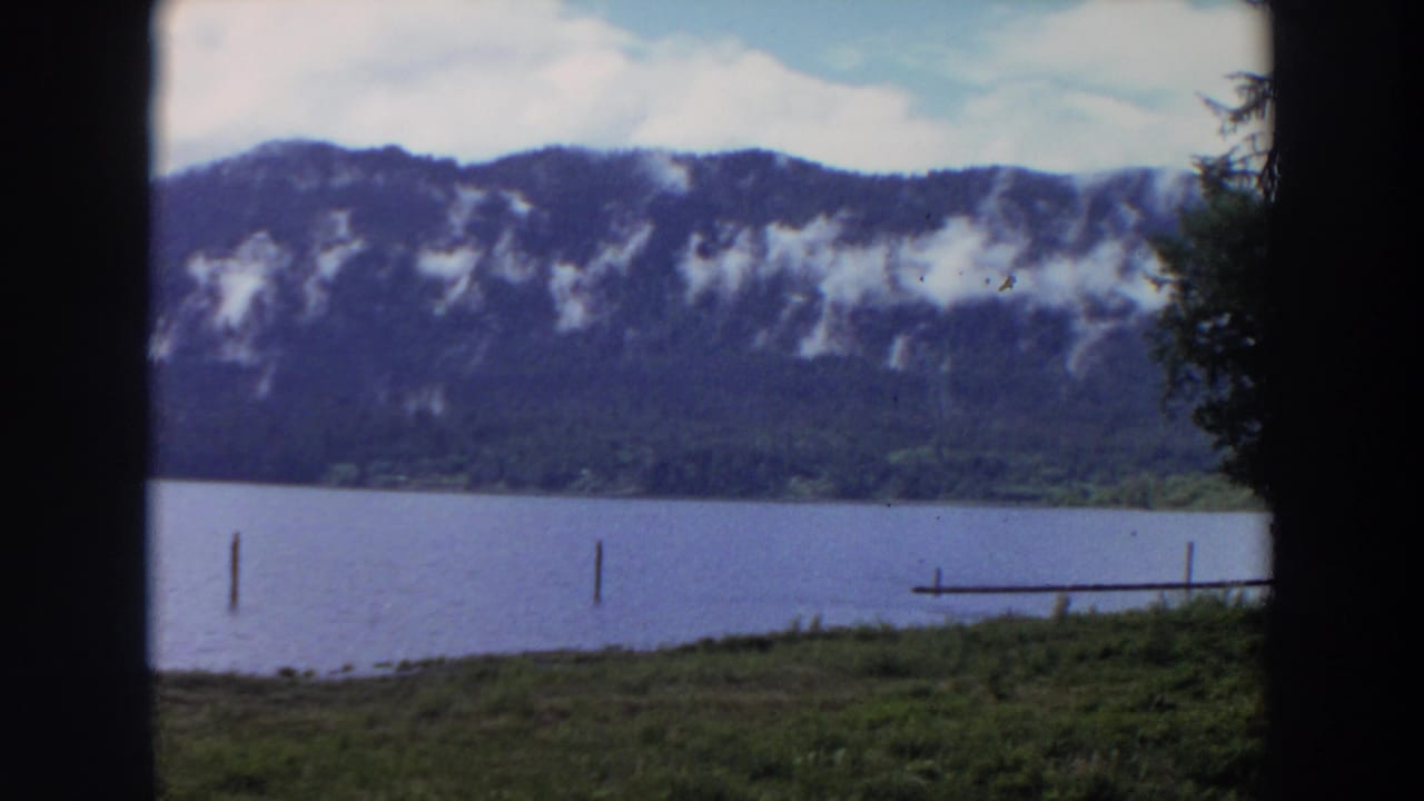 1964 California 8mm frame of a lake below a wooded mountain ridge, with posts in the water and a dock at the right edge.