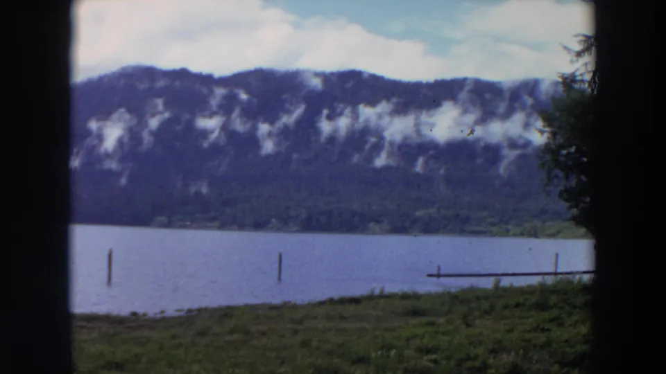 1964 California 8mm frame of a lake below a wooded mountain ridge, with posts in the water and a dock at the right edge.