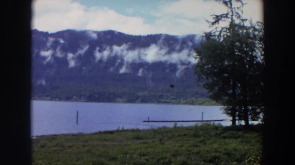 1964 California home-movie frame with a tree beside a lake, a small dock, and clouds along a forested mountain ridge.