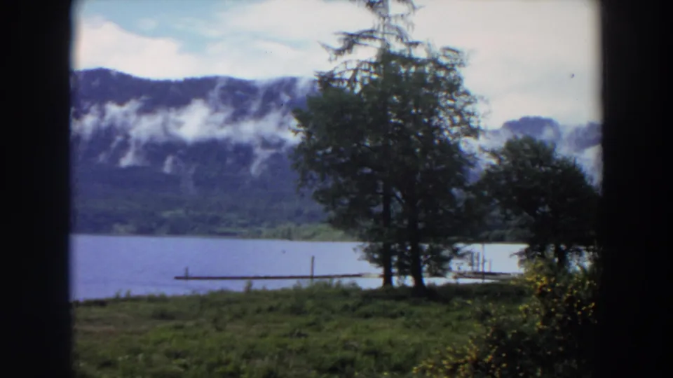 1964 California 8mm frame showing lakeside trees, grass, a narrow dock, and mist along distant mountain slopes.
