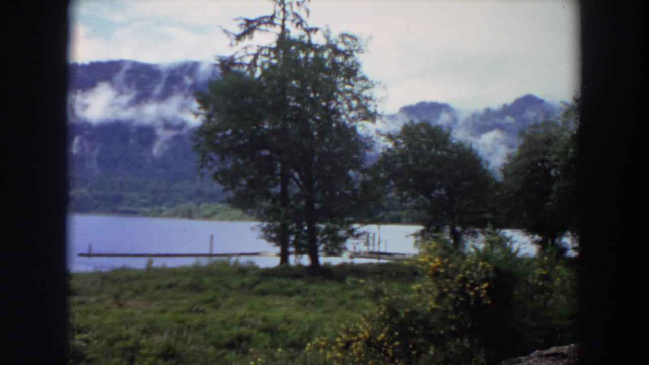 1964 California home-movie still with trees, shoreline grass, lake water, dock posts, and low mist on mountain ridges.