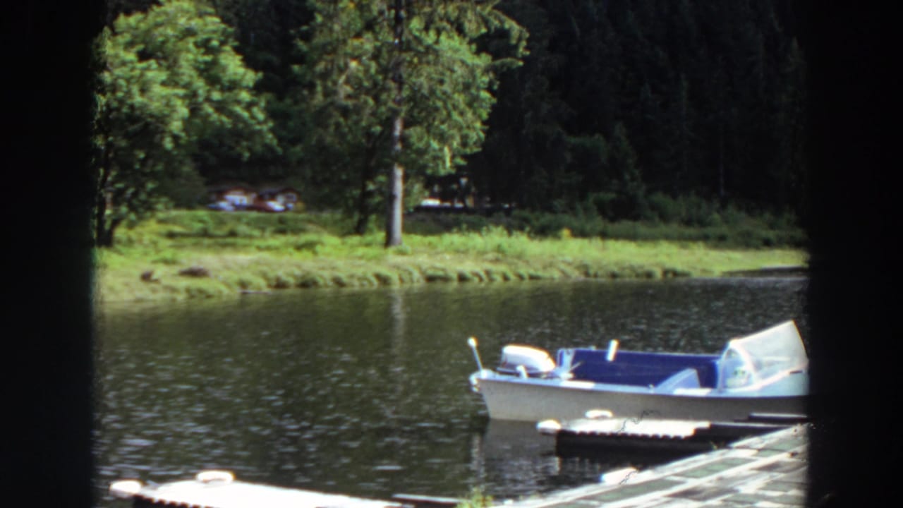 Empty lakeside motorboat in a 1964 California home-movie frame, with a dock edge, rippled water, grass, and trees visible.