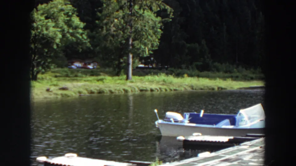 1964 California 8mm view of an empty white and blue motorboat at a dock, with calm water and wooded shoreline behind it.