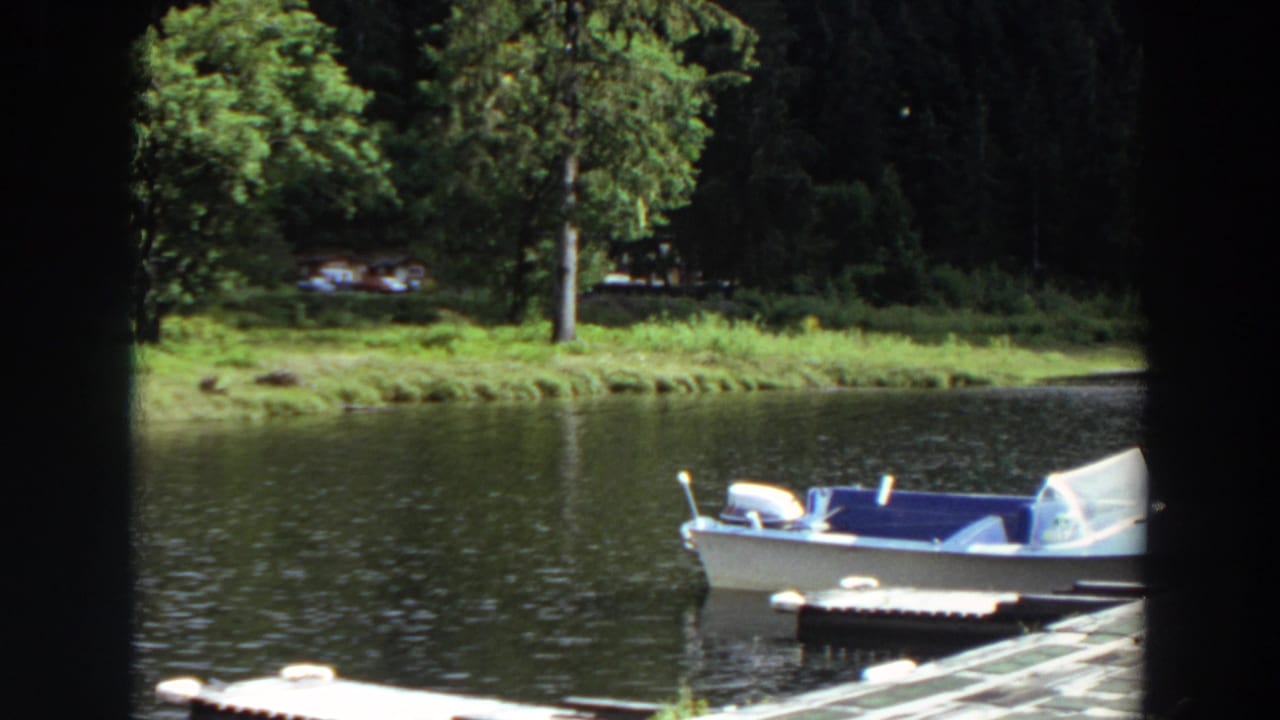 White motorboat moored near a wooden dock on a calm lake with trees and cabins across the water.
