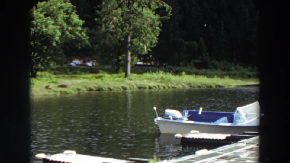 1964 California 8mm frame showing a white motorboat with blue seats beside a dock, with grassy shoreline, trees, cabins, and parked cars beyond the lake.