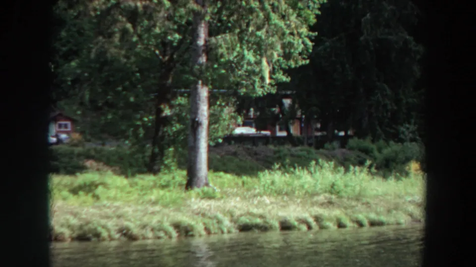 1964 California 8mm frame looking across calm lake water toward a grassy shoreline with tall trees, small cabins, and parked cars in shade.